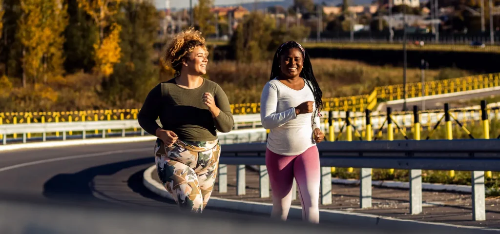 Two women jogging on a sunny road near a park with trees and distant buildings in the background.