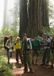 A group of six hikers with backpacks stand on a forest trail, surrounded by tall trees.