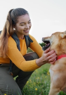 A person in a yellow sweater and backpack kneels in a field, smiling and petting a brown dog with a red collar.