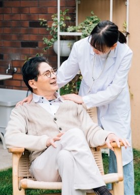 A person in a white coat stands behind a seated man in a light sweater and glasses, outdoors, with hands on his shoulders. They are smiling at each other.