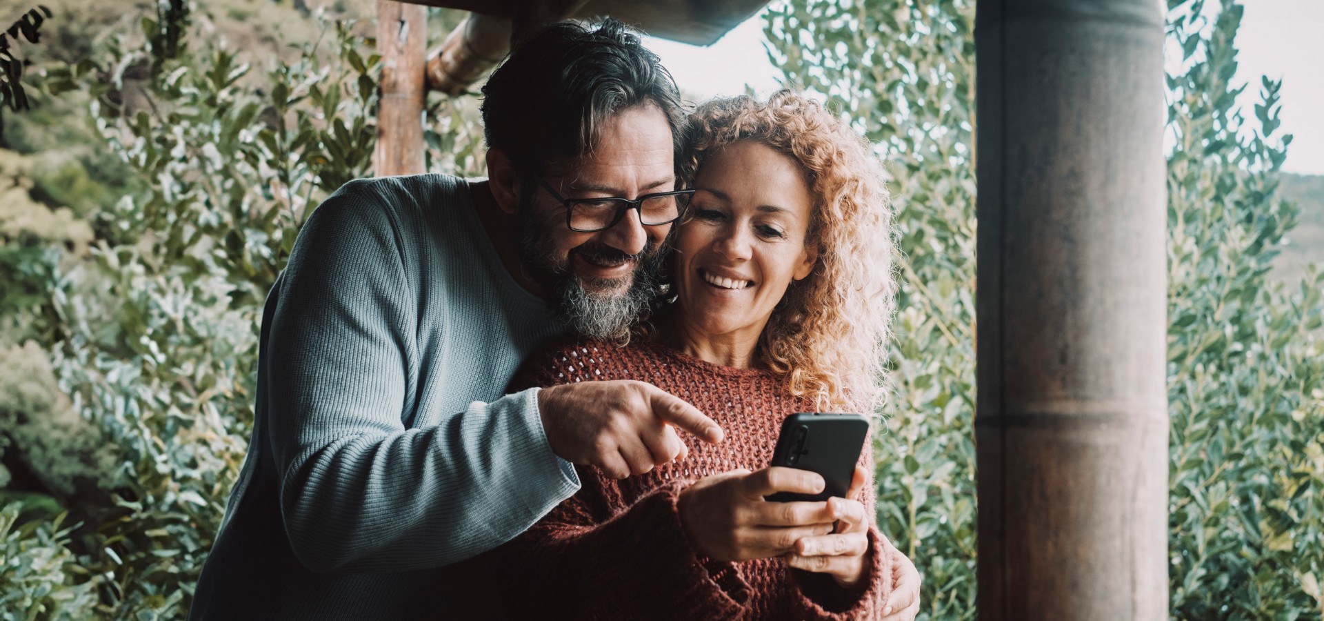 A couple stands outdoors, smiling and looking at a phone together.
