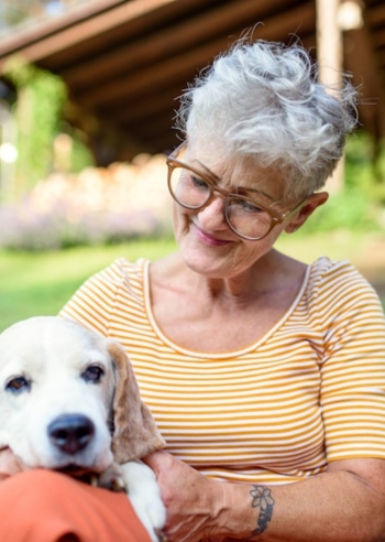 Elderly woman with glasses and short gray hair smiles while holding a white and tan dog outdoors.