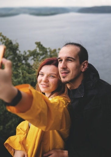 A couple in a yellow and black raincoat takes a selfie with a scenic lake and forest in the background.