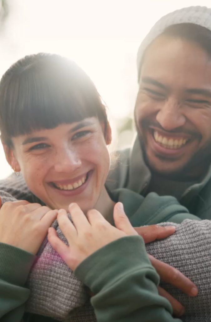 A couple smiling and embracing, with the sun shining behind them. Both are wearing casual clothing.