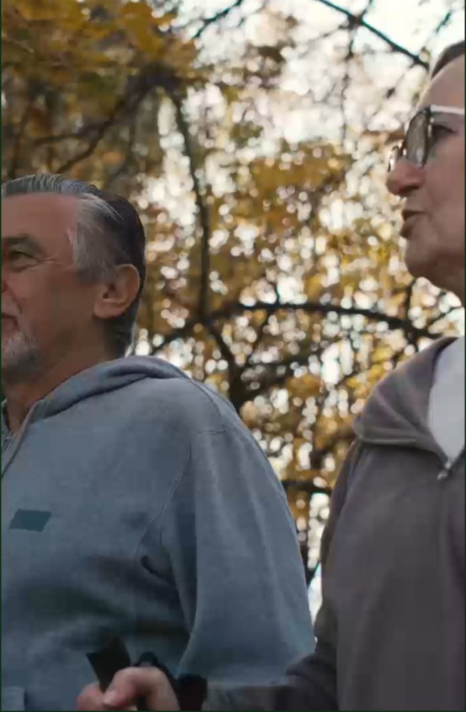 Two men walking outdoors under a canopy of autumn leaves, both wearing glasses and casual jackets.