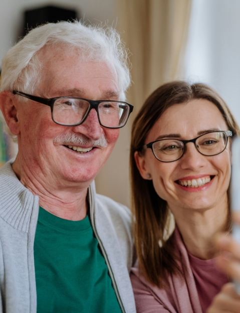 An older man and a younger woman, both wearing glasses, smile while taking a selfie indoors.