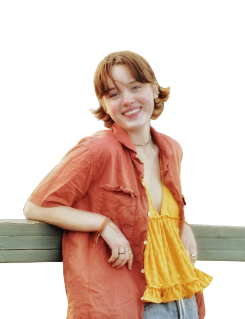 A person with short hair smiles, leaning against a wooden railing while wearing an orange shirt over a yellow top.