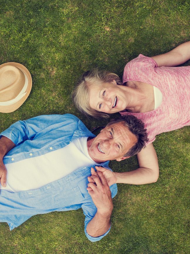 A man and woman lie on grass smiling, with a hat beside them.