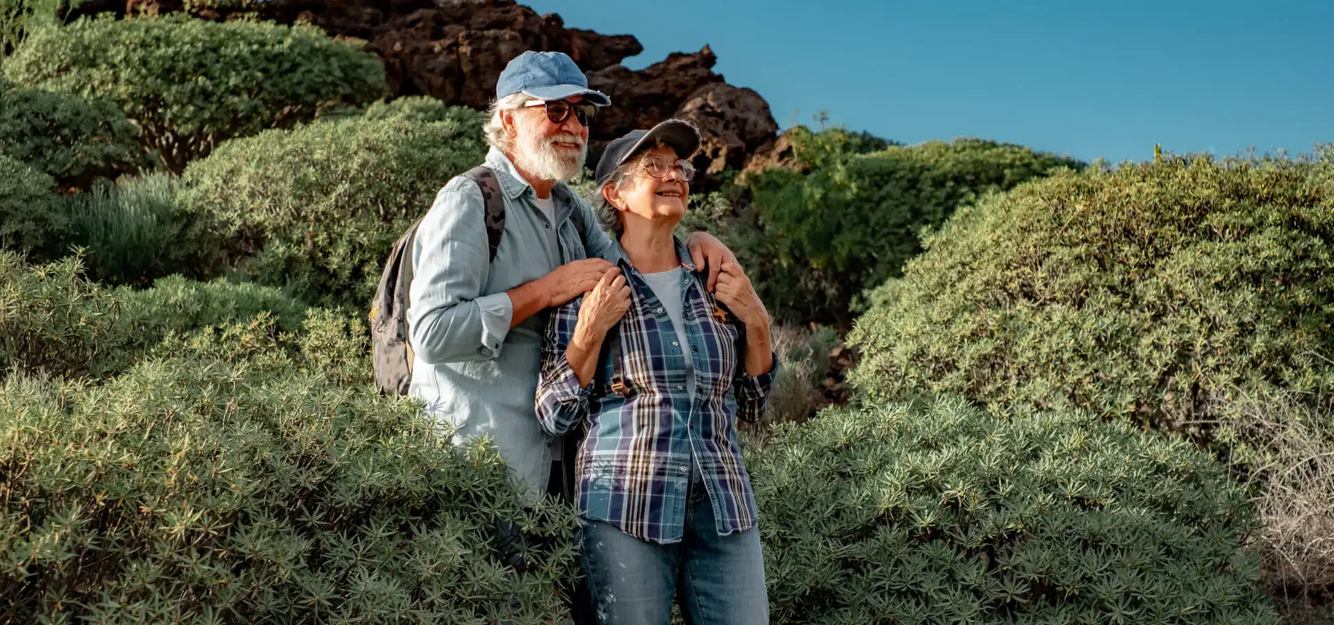 Older couple in casual clothes and hats hiking through lush greenery, smiling and enjoying the scenery.