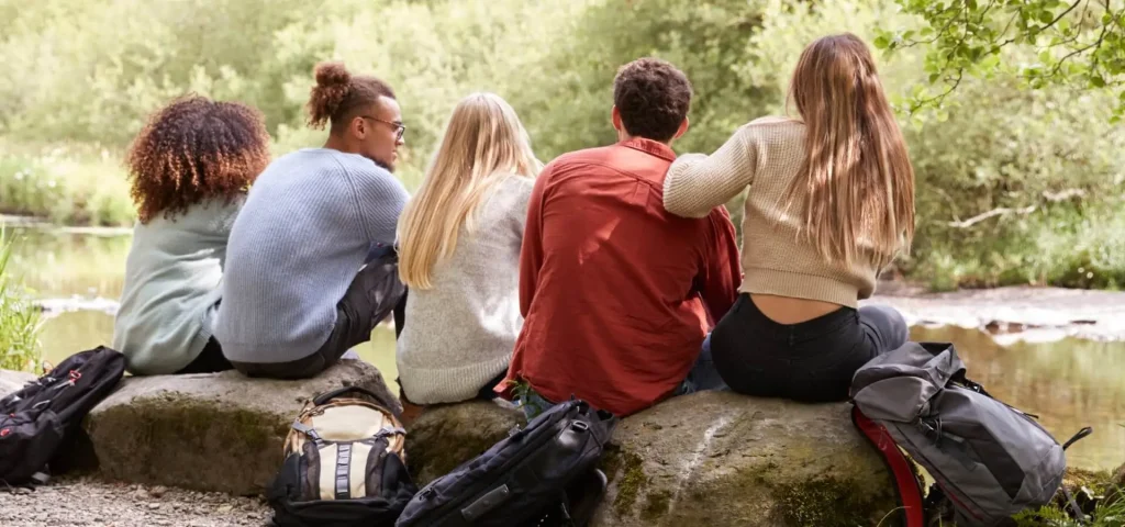 Five people sit on a rock by a riverside, with backpacks nearby, looking away towards the water and trees.