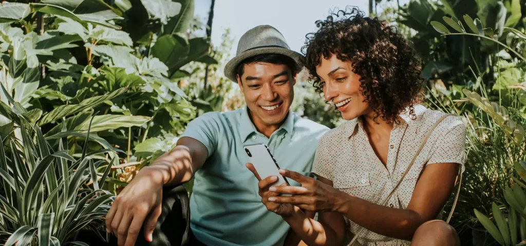A man and woman sitting outdoors, smiling and looking at a smartphone together, surrounded by greenery.