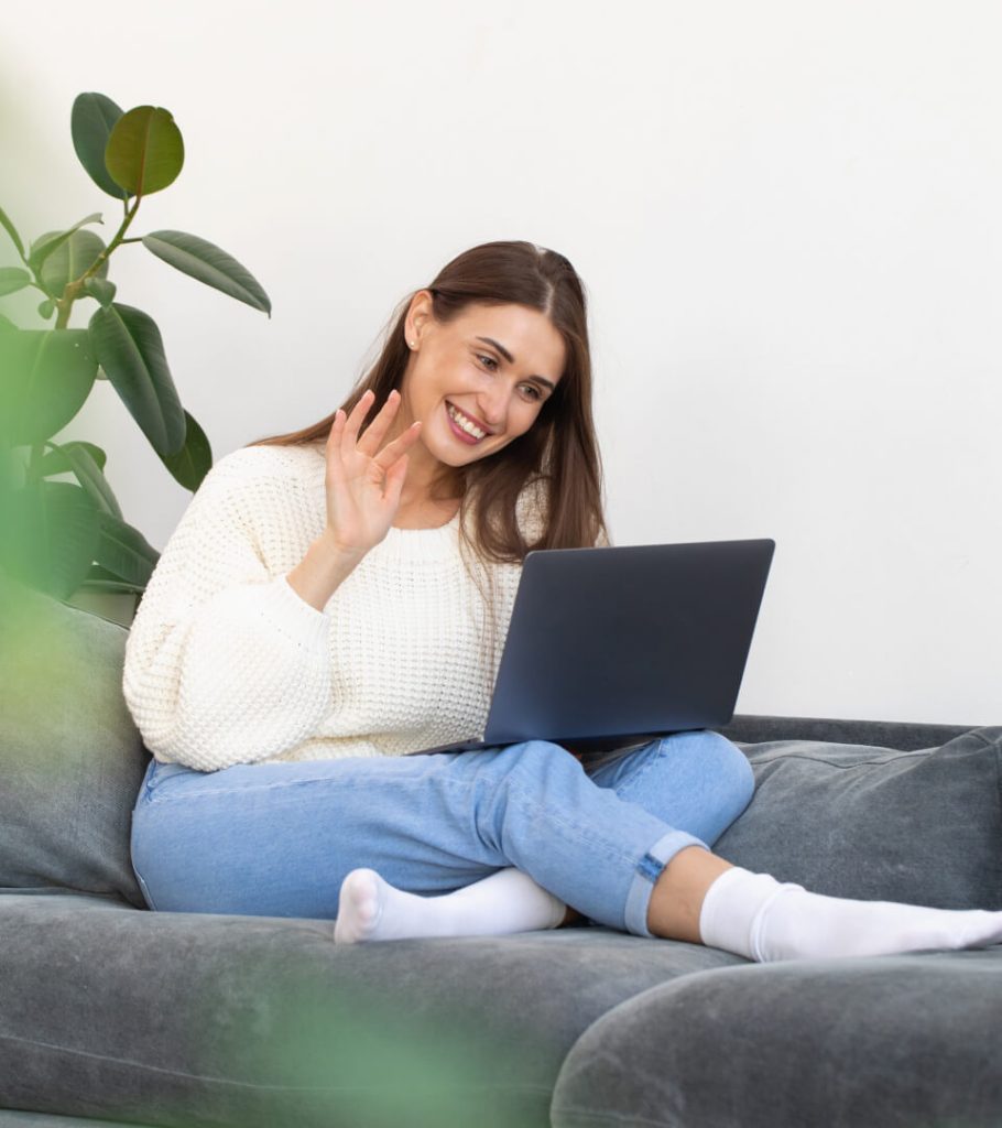A woman sitting on a couch waves at her laptop screen, appearing engaged in a video call. She is wearing a white sweater and jeans, with a green plant in the background.