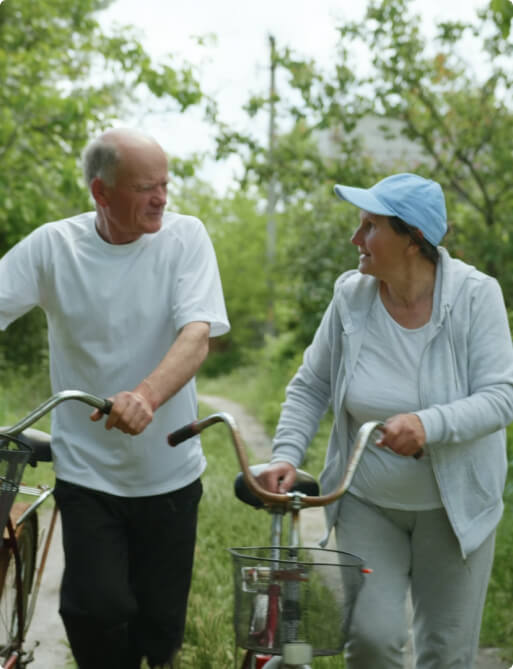 An elderly couple walks with bicycles along a path surrounded by greenery, engaged in conversation.