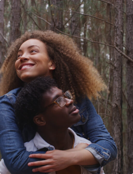 A woman with curly hair smiles while piggybacking on a man wearing glasses in a forest setting.