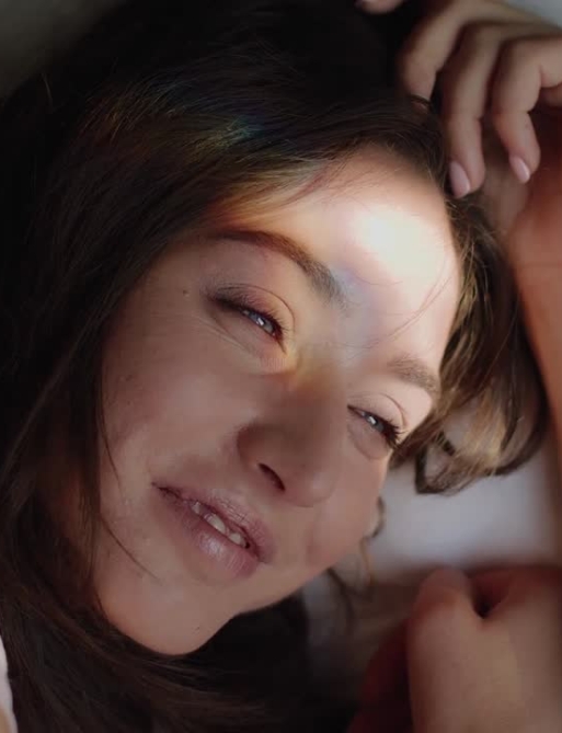 Close-up of a person lying down, smiling slightly, with light and shadow on their face. Their hand rests near their head.
