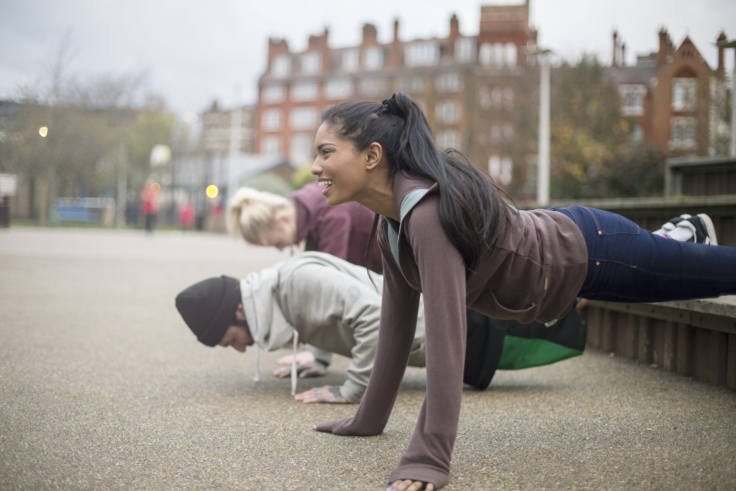 Three people outdoors, following sample routines, are doing push-ups on a pavement with buildings in the background.