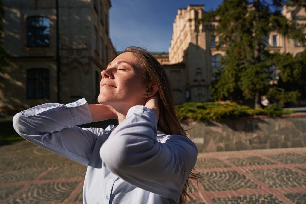 A woman stands outdoors with her eyes closed, basking in the sun's warmth. She gently touches her neck with both hands, embodying tranquility. A building and trees frame this serene scene, making it a perfect sample post for relaxation and mindfulness.