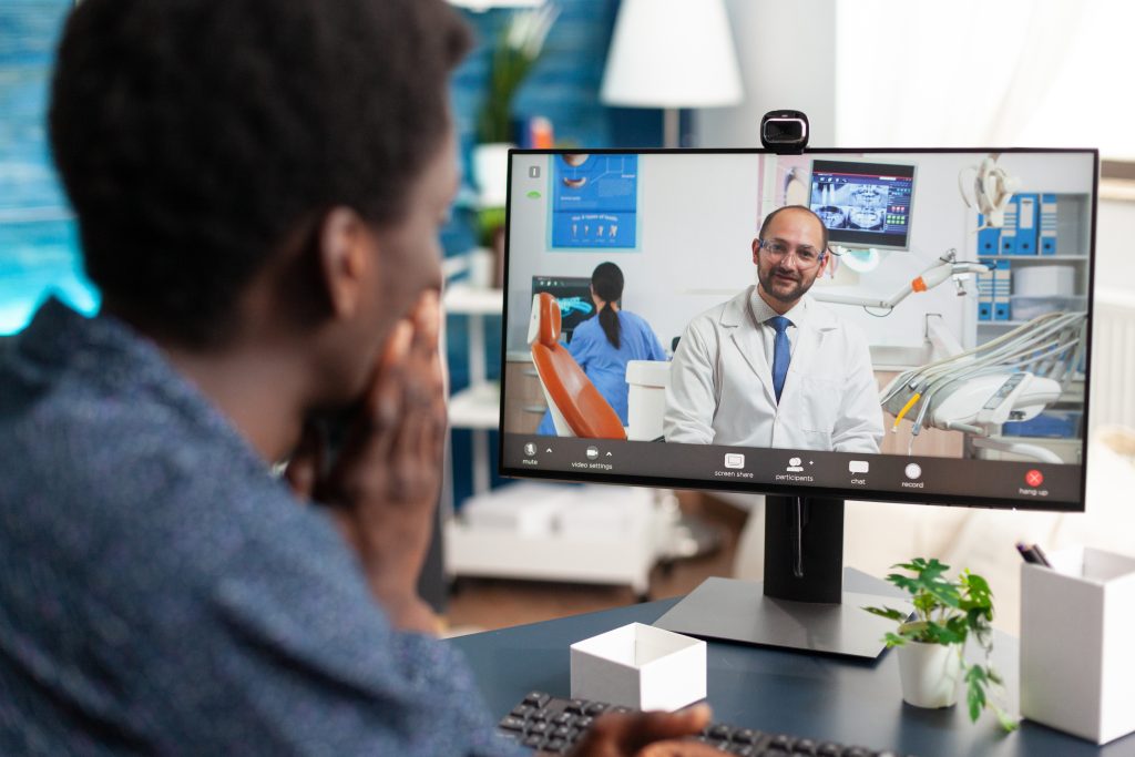 During a healthcare consultation, a person participates in a video call with a doctor displayed on a computer screen, surrounded by dental equipment.
