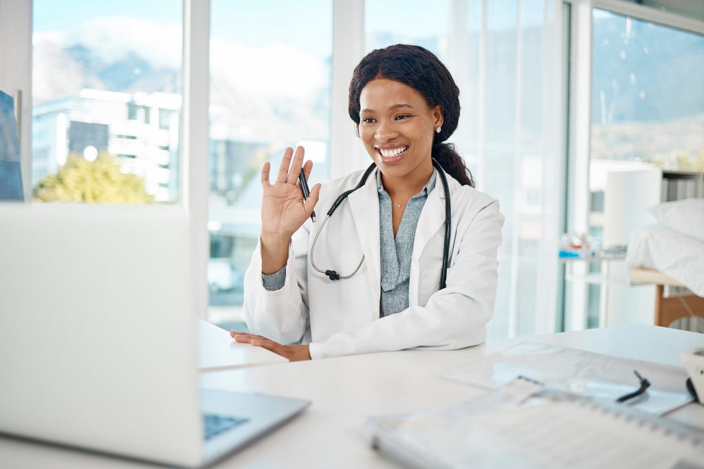 The doctor in a white coat, smiling and waving at a laptop during a healthcare consultation, is seated at a desk with medical documents in a bright office with large windows.